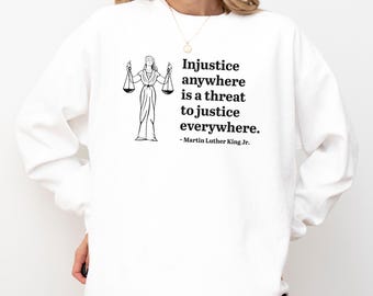 Op de afbeelding: Witte sweatshirt met een zwart-wit illustratie van Vrouwe Justitia met weegschalen en de quote "Onrechtvaardigheid waar dan ook is een bedreiging voor gerechtigheid overal" van Martin Luther King Jr. Een comfortabele, casual stijl.