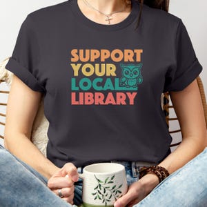 May include: Dark gray t-shirt with the text "SUPPORT YOUR LOCAL LIBRARY" in orange, yellow, teal, and pink. A teal owl graphic is next to the word "LIBRARY". The shirt is being held by a person.
