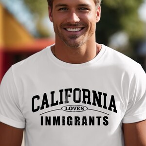 Puede incluir: Camiseta blanca con el texto negro "CALIFORNIA LOVES IMMIGRANTS". La camiseta también tiene el texto "ONLY THE DESIGN IS FOR SALE". La persona de la imagen está sonriendo.