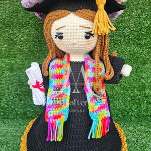 May include: Crocheted graduation doll wearing a black cap and gown with a colorful scarf. The doll is holding a white scroll and has a gold tassel on the cap. The doll is sitting on a green surface.