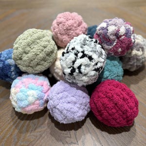 May include: A collection of handmade, textured yarn balls in various colors, including pink, blue, green, purple, and white. The balls are arranged in a pile, showcasing their soft, fluffy texture. These decorative balls are perfect for crafts.