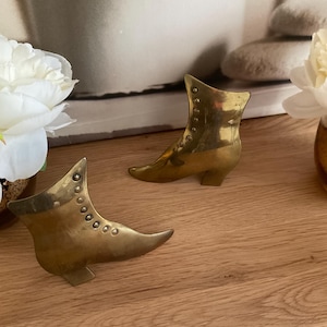 Puede incluir: Dos sujetalibros de metal dorado en forma de bota. Los sujetalibros están detallados con una serie de pequeños círculos a lo largo de la parte superior de la bota.