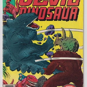 May include: A vintage comic book cover featuring the title "Devil Dinosaur" in large red letters against a yellow background. The cover depicts a battle scene with dinosaurs and the text "Marvel Comics Group".