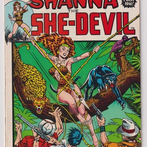 May include: Vintage comic book cover of "Shanna the She-Devil" from Marvel Comics. The cover features a woman in a leopard print outfit, swinging on a vine, with a jaguar and panther. Text includes "And a Jungle Queen is Born!"
