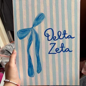 Puede incluir: Una pintura sobre lienzo con un lazo azul y las palabras "Delta Zeta" en escritura azul. El fondo presenta rayas verticales en azul claro y blanco roto. La pintura es sostenida por una persona.