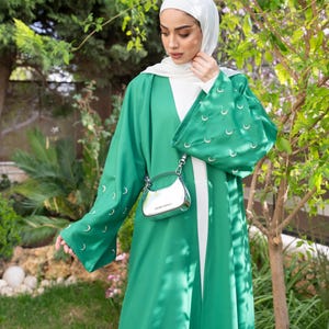 May include: A vibrant green open-front abaya featuring wide sleeves adorned with small silver crescent moon embellishments. The garment is worn over a plain white under-layer and paired with a white hijab, complemented by a small metallic silver handbag.