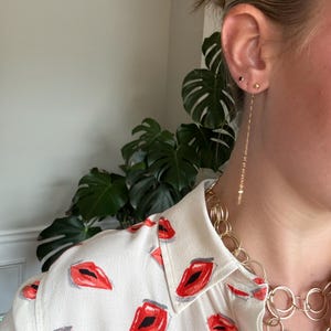 May include: A close-up of a woman's ear with two small gold stud earrings and a long, delicate gold chain earring. The woman is wearing a white blouse with a red lip print and a gold chain necklace.