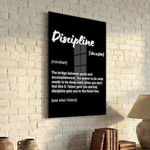May include: A black framed wall art print with the word "Discipline" in white script. The definition of discipline is provided below the word, along with its pronunciation and the phrase "(see also: Vision)." The print is displayed on a white brick wall.