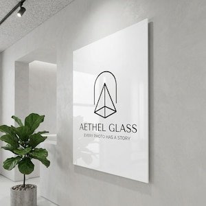 Peut inclure: Un grand panneau rectangulaire blanc avec le texte "Aethel Glass" et un logo géométrique. Le texte "Every photo has a story" est en dessous. Le panneau est fixé à un mur gris clair dans un intérieur moderne.