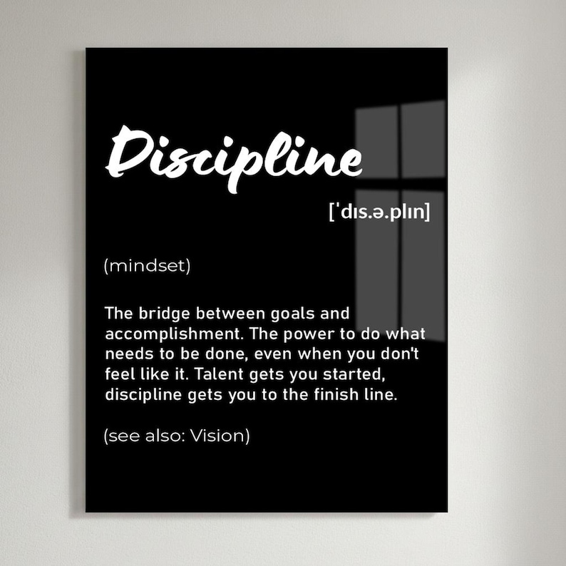 May include: A black framed wall art print with the word "Discipline" in white script. The definition of discipline is provided below the word, along with its pronunciation and the phrase "(see also: Vision)." The print is displayed on a white brick wall.