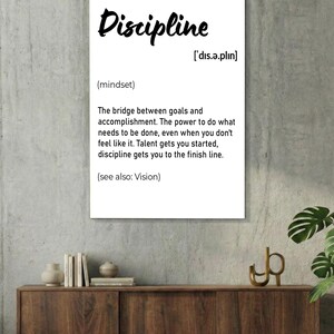 May include: A black framed wall art print with the word "Discipline" in white script. The definition of discipline is provided below the word, along with its pronunciation and the phrase "(see also: Vision)." The print is displayed on a white brick wall.