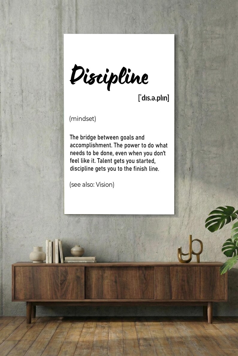 May include: A black framed wall art print with the word "Discipline" in white script. The definition of discipline is provided below the word, along with its pronunciation and the phrase "(see also: Vision)." The print is displayed on a white brick wall.