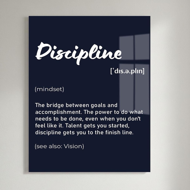May include: A black framed wall art print with the word "Discipline" in white script. The definition of discipline is provided below the word, along with its pronunciation and the phrase "(see also: Vision)." The print is displayed on a white brick wall.