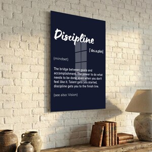 May include: A black framed wall art print with the word "Discipline" in white script. The definition of discipline is provided below the word, along with its pronunciation and the phrase "(see also: Vision)." The print is displayed on a white brick wall.