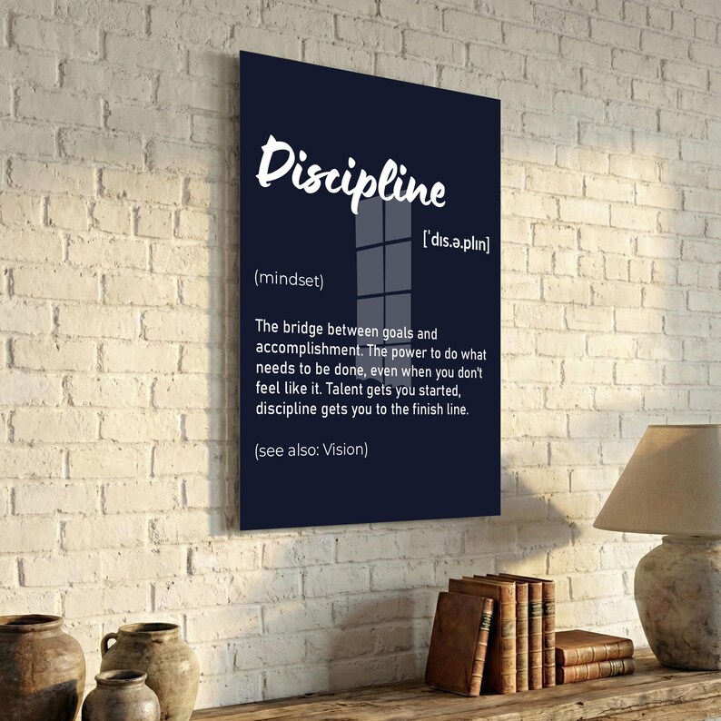 May include: A black framed wall art print with the word "Discipline" in white script. The definition of discipline is provided below the word, along with its pronunciation and the phrase "(see also: Vision)." The print is displayed on a white brick wall.