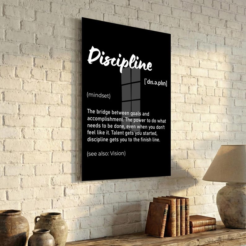 May include: A black framed wall art print with the word "Discipline" in white script. The definition of discipline is provided below the word, along with its pronunciation and the phrase "(see also: Vision)." The print is displayed on a white brick wall.