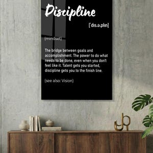 May include: A black framed wall art print with the word "Discipline" in white script. The definition of discipline is provided below the word, along with its pronunciation and the phrase "(see also: Vision)." The print is displayed on a white brick wall.