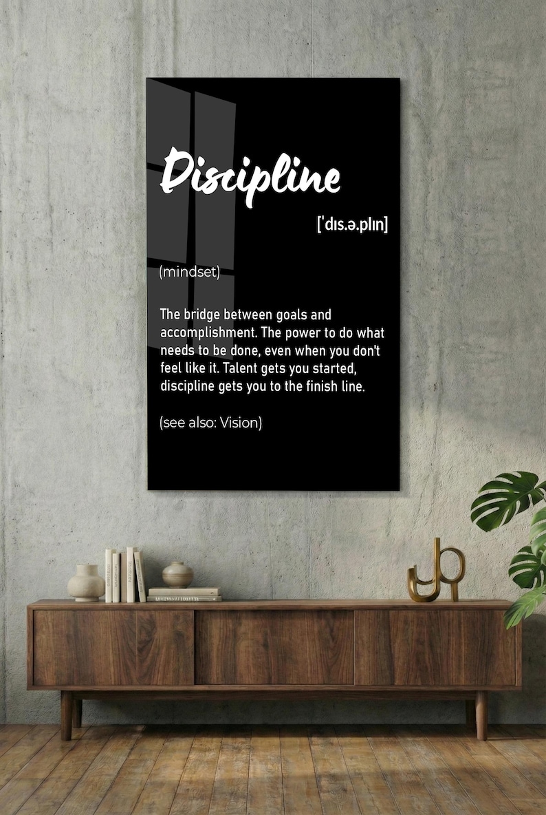 May include: A black framed wall art print with the word "Discipline" in white script. The definition of discipline is provided below the word, along with its pronunciation and the phrase "(see also: Vision)." The print is displayed on a white brick wall.