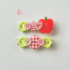 May include: Two decorative hair clips. One clip features a red apple, pink buttons, and a heart-shaped pearl accent. The other clip has a gingham button, pink buttons, and a heart-shaped pearl accent. Both clips have green flower-shaped accents.