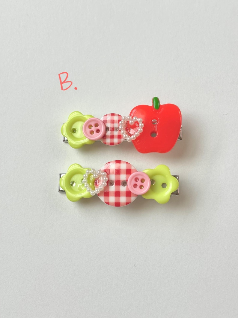 May include: Two decorative hair clips. One clip features a red apple, pink buttons, and a heart-shaped pearl accent. The other clip has a gingham button, pink buttons, and a heart-shaped pearl accent. Both clips have green flower-shaped accents.