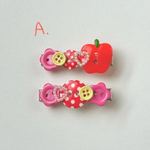 May include: Two decorative hair clips. One clip features a red apple, a polka dot button, a heart-shaped pearl design, and a pink flower button. The other clip has a polka dot button, a heart-shaped pearl design, and a pink flower button.