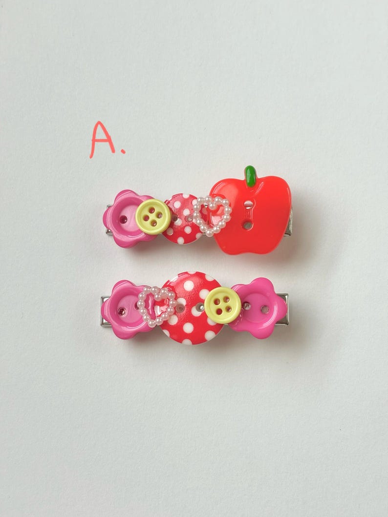 May include: Two decorative hair clips. One clip features a red apple, a polka dot button, a heart-shaped pearl design, and a pink flower button. The other clip has a polka dot button, a heart-shaped pearl design, and a pink flower button.