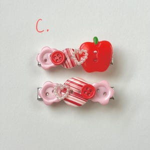 May include: Two decorative hair clips. Each clip features a combination of pink flower-shaped buttons, red and white striped elements, red buttons, and a heart-shaped pearl accent. One clip has a red apple charm.