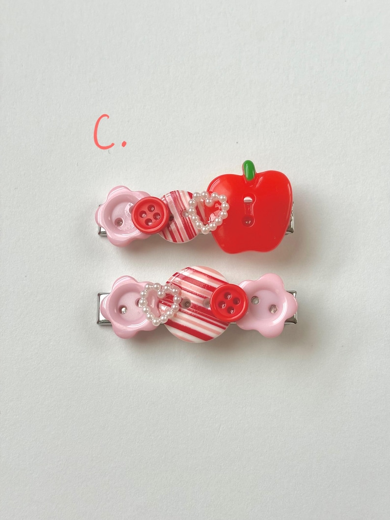 May include: Two decorative hair clips. Each clip features a combination of pink flower-shaped buttons, red and white striped elements, red buttons, and a heart-shaped pearl accent. One clip has a red apple charm.