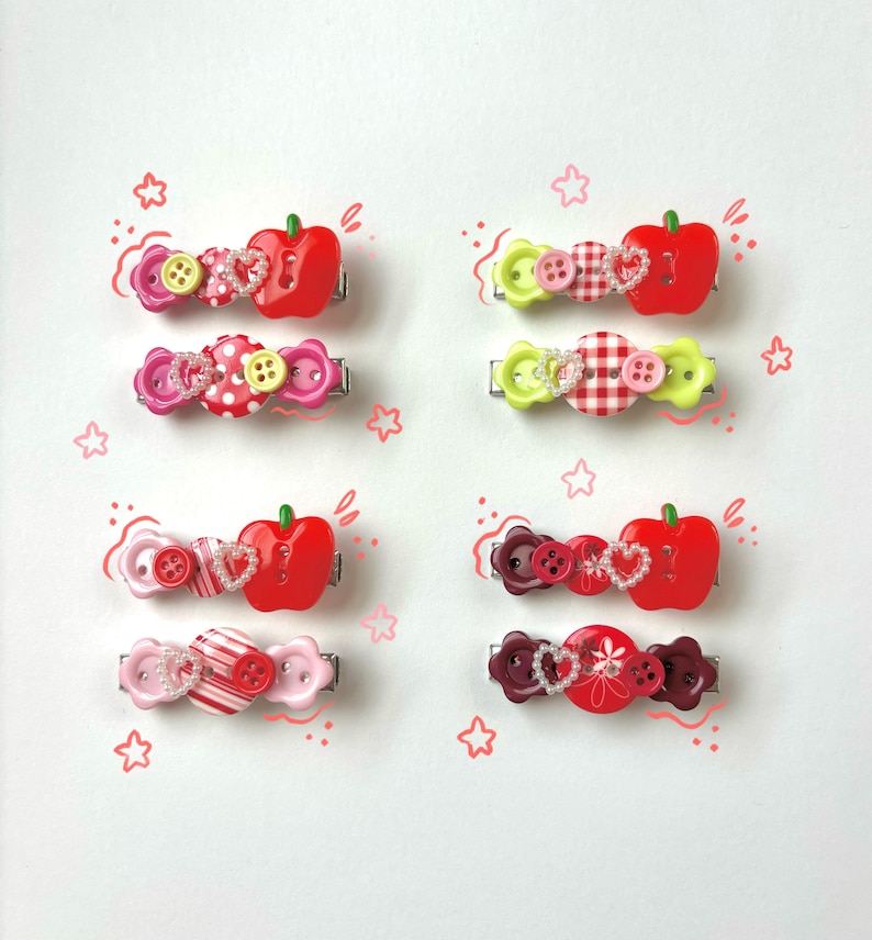 May include: A collection of colorful hair clips with various designs. Each clip features a combination of buttons, heart-shaped embellishments, and a red apple-shaped accent. The clips are in shades of pink, red, and green, with a mix of patterns.