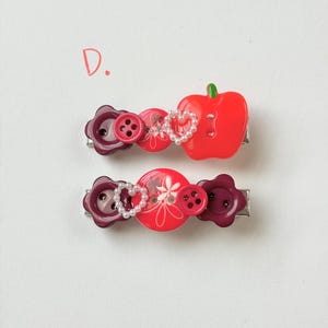 May include: Two decorative hair barrettes. One features a red apple, the other has a heart-shaped pearl design. Both have red and purple button accents and are on silver metal clips. The letter "D" is in the upper left corner.