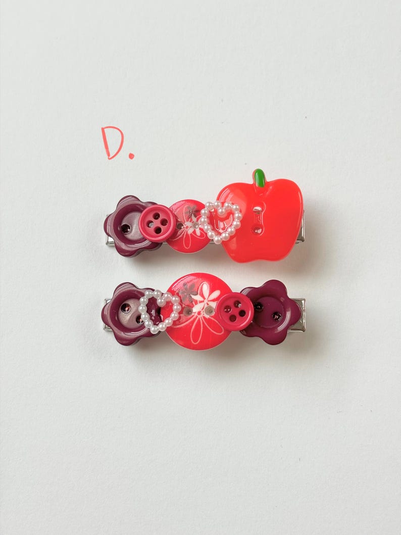 May include: Two decorative hair barrettes. One features a red apple, the other has a heart-shaped pearl design. Both have red and purple button accents and are on silver metal clips. The letter "D" is in the upper left corner.
