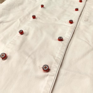 Può includere: Giacca da chef bianca con bottoni rossi. I bottoni hanno un design a croce bianca.