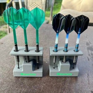 Könnte beinhalten: Zwei graue Dart-Halter mit dem Namen Maurizio auf der Vorderseite. Ein Halter hat drei grüne Darts und der andere drei blaue und schwarze Darts.