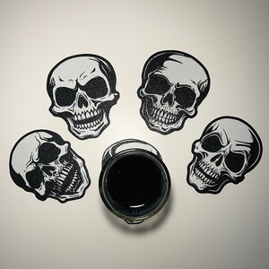 Könnte beinhalten: Vier schwarze und weiße Totenkopf-Untersetzer mit einem schwarzen Glas in der Mitte. Die Untersetzer sind aus einem dicken, strapazierfähigen Material gefertigt und haben ein detailliertes Totenkopf-Design.
