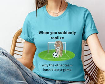 Op de afbeelding: Een lichtblauw t-shirt met een afbeelding van een tijger op een voetbalveld. De tekst op het shirt luidt "Wanneer je je plotseling realiseert waarom het andere team geen wedstrijd heeft verloren".