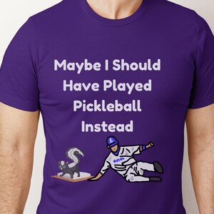 Puede incluir: Camiseta morada con texto blanco que dice "Maybe I Should Have Played Pickleball Instead". Un jugador de béisbol de dibujos animados se desliza hacia el plato de home, con una mofeta detrás de él.