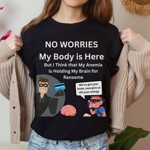 Può includere: Maglietta nera con una grafica divertente di un personaggio dei cartoni animati che tiene una carta di credito e il testo "NO WORRIES My Body is Here But I Think that My Anemia is Holding My Brain for Ransome We've got your brain, now give us all your energy".