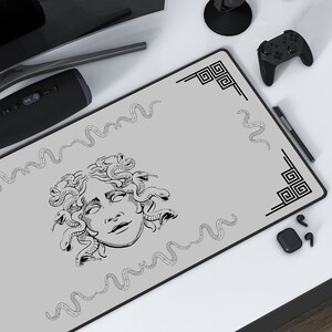 May include: A rectangular grey desk mat with a black and white illustration of Medusa's face, surrounded by snakes and decorative borders. The mat is on a white desk with a keyboard, mouse, and other tech accessories.