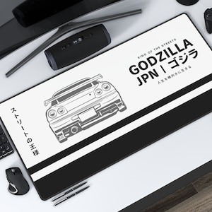 Pode incluir: Tapete de secretária retangular branco e preto com um gráfico de carro e texto japonês. O tapete exibe as palavras "GODZILLA JPN" e "KING OF THE STREETS". O gráfico do carro é uma ilustração detalhada de um carro desportivo.