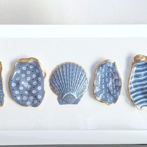 May include: A rectangular white shadow box frame containing five decorative seashells arranged in a horizontal row. Each shell is painted in shades of blue and white with distinct patterns including dots, stripes, and lines, all featuring metallic gold edges.