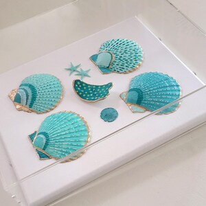 May include: A collection of painted seashells in shades of turquoise and gold, arranged on a white surface within a clear display case. The seashells vary in size and shape, with intricate patterns and gold accents. The arrangement includes starfish and a small, round shell.