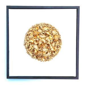 May include: A square wall art piece featuring a circular arrangement of various gold-painted seashells. The collection of shells is densely packed into a round cluster, centered on a plain white background and enclosed within a thin black frame.