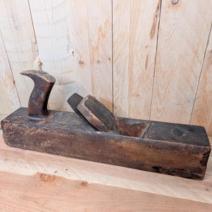 May include: A vintage wooden hand plane with a dark, weathered appearance. The plane features a long, rectangular body with a curved handle and a blade. The wood shows signs of age and use, with visible wear and tear.