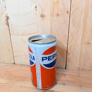 May include: Vintage Pepsi Cola can with a white, red, and blue design. The can has a classic logo and is slightly dented, showing signs of age. The can is cylindrical and made of metal.