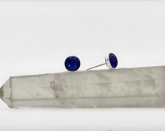 May include: A pair of stud earrings with a round, deep blue, glittery design set in a silver bezel. One earring is lying on a clear, rectangular crystal. The other earring is standing upright.
