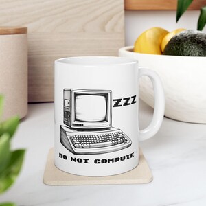 May include: White ceramic mug featuring a black and white illustration of a vintage computer with the word "zzz" and the phrase "DO NOT COMPUTE". The mug has a curved handle and is sitting on a square coaster.