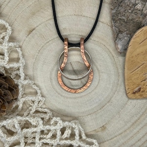 Könnte beinhalten: Eine Kupfer- und Silber-Anhänger-Halskette mit schwarzem Band. Der Anhänger hat zwei ineinandergreifende Ringe, einen kupferfarbenen mit strukturiertem Muster und einen silberfarbenen. Die Halskette wird auf einer Holzoberfläche präsentiert.