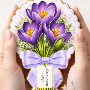 May include: A decorated cookie shaped like a bouquet of purple crocus flowers with yellow centers and green stems. The cookie has a light purple bow and a tag that says "Happy Women's Day!"
