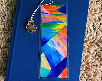 May include: A colorful bookmark with a geometric design of blue, orange, and green hues. The bookmark is attached to a gold-colored charm with a sun design and a blue center, resting on a closed blue book.