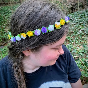 May include: A floral crown featuring small, colorful fabric flowers in yellow, white, and purple, set against a green base. The flowers are arranged in a circular pattern. The crown is worn on a person's head, with their hair visible.